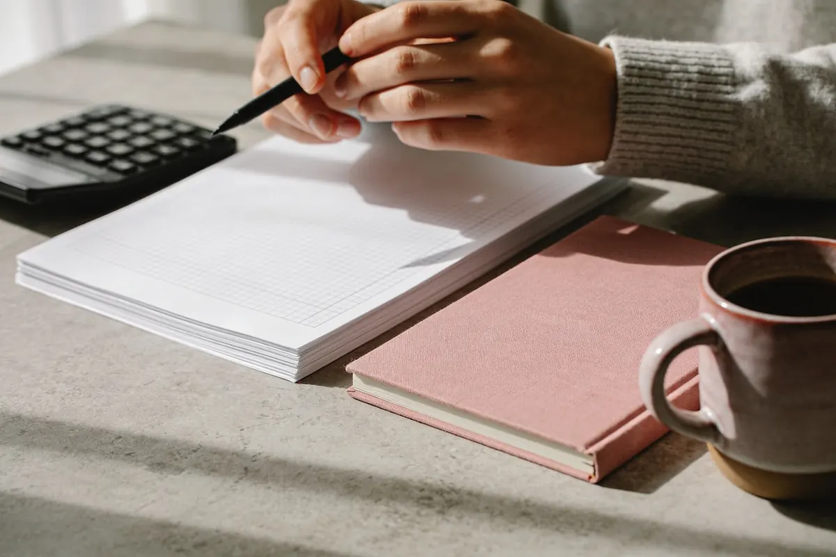Hände mit Kugelschreiber neben Tabellen, Taschenrechner und rosa Notizbuch auf einem Steintisch